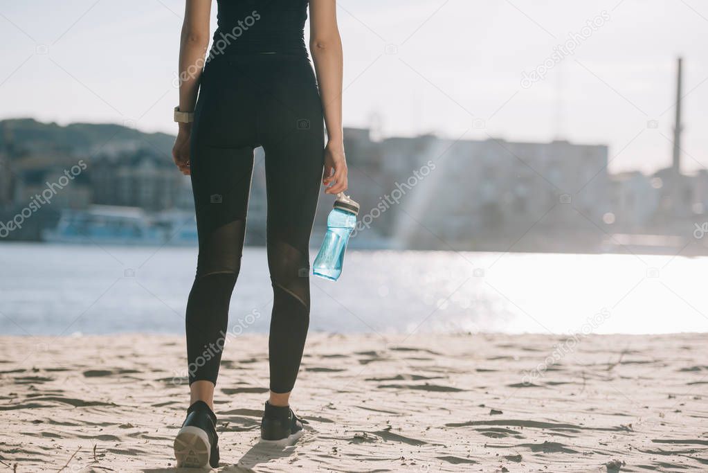 cropped view of sportswoman with water in bottle walking on beach with back light