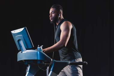 handsome young african american man in earphones exercising on treadmill and looking away clipart