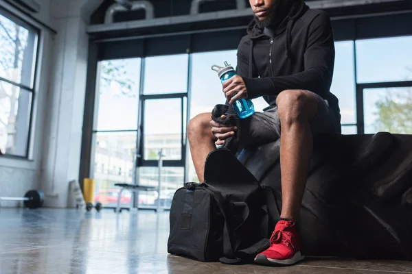 年轻的非洲裔美国运动员在健身房坐轮胎时手持瓶装水和毛巾的裁剪镜头 — 图库照片