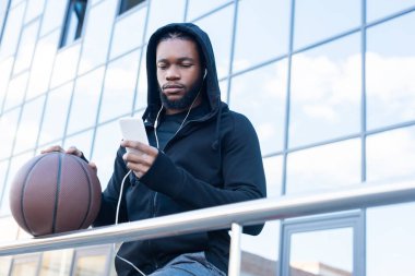 sokak basketbol topuyla dururken smartphone kullanarak kulaklık sportif Afro-Amerikan insanın düşük açılı görünüş