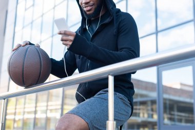 Afrika kökenli Amerikalı adam sokakta basketbol topu tutarken smartphone kullanarak kulaklık gülümseyen in kırpılmış atış