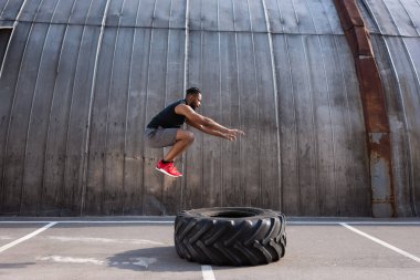 muscular african american sportsman jumping while training with tyre on street  clipart