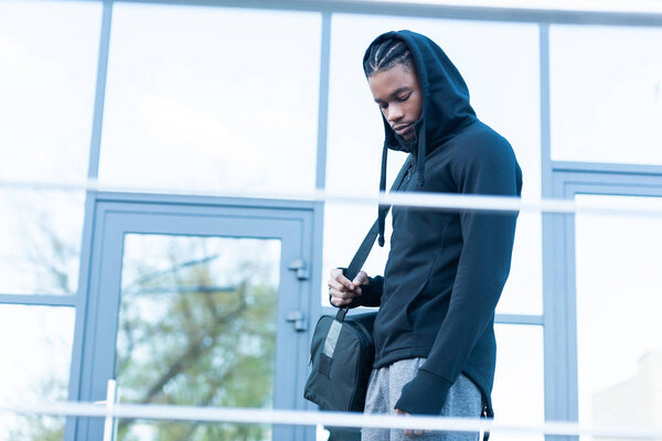 Вид сбоку молодого афроамериканца, который держит спортивную сумку и смотрит вниз на улице
