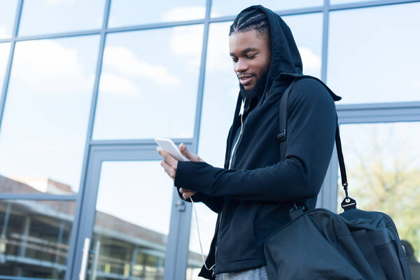 улыбающийся молодой африканский американец со спортивной сумкой с помощью смартфона на улице
