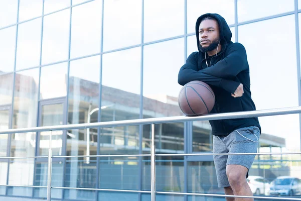 pensive african american man in earphones leaning at railings with basketball ball and looking away