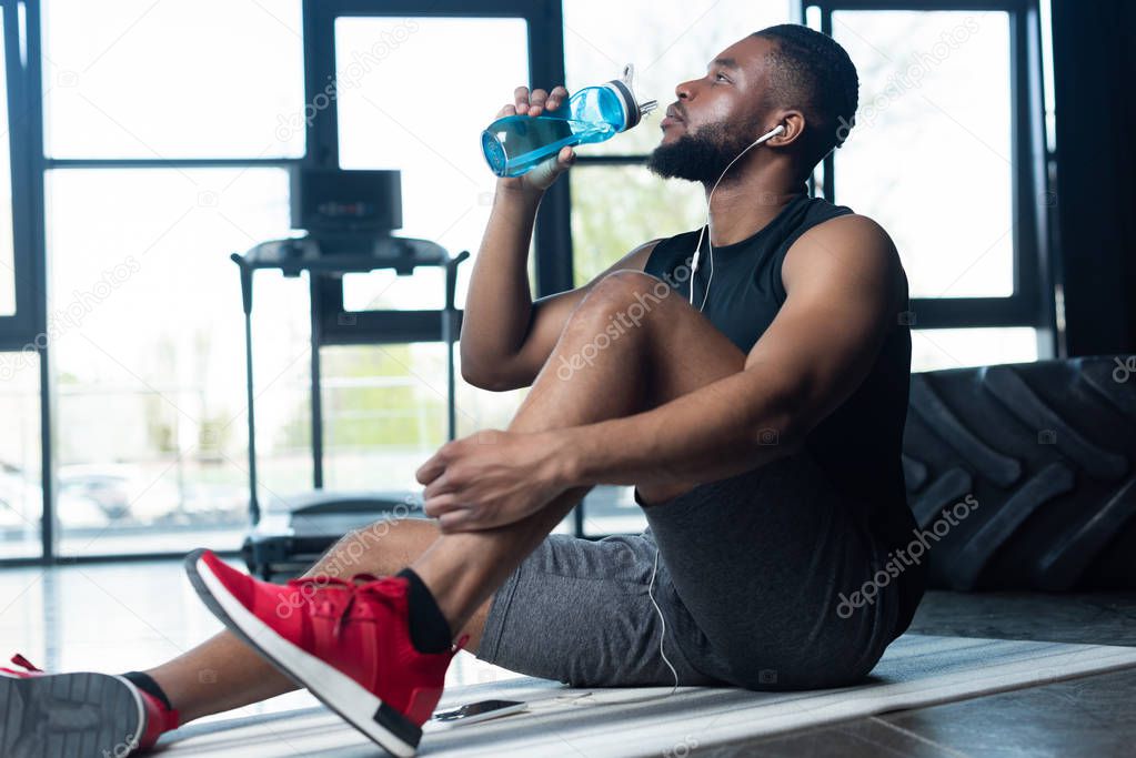 young african american sportsman in earphones sitting on yoga mat and drinking water in gym