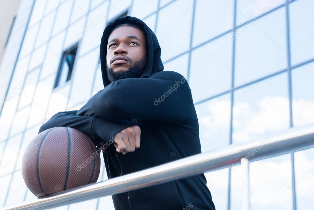 low angle view of pensive african american sportsman looking away while standing with basketball ball on street