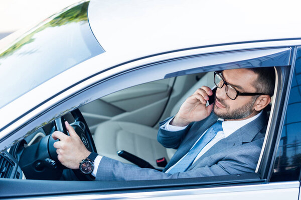 side view of businessman in eyeglasses talking on smartphone while driving car 