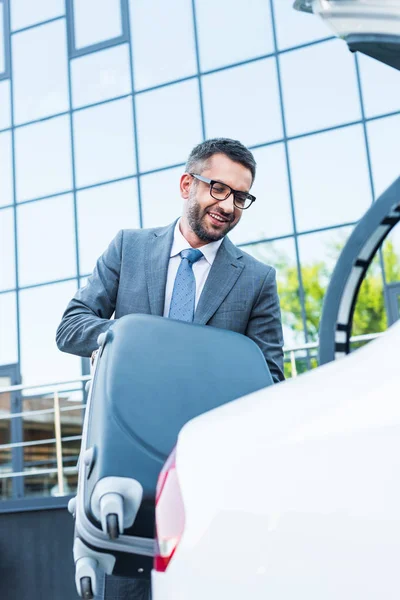 portrait of businessman in eyeglasses putting luggage into car on parking