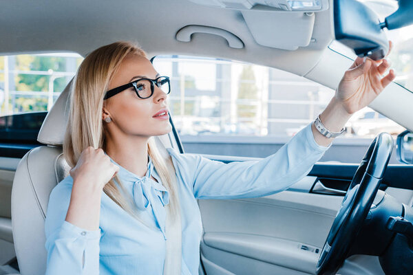 Вид сбоку блондинки в очках, смотрящей в зеркало заднего вида в машине
