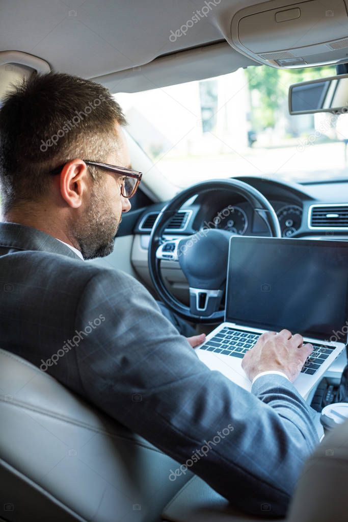 side view of businessman in eyeglasses using laptop with blank screen in car