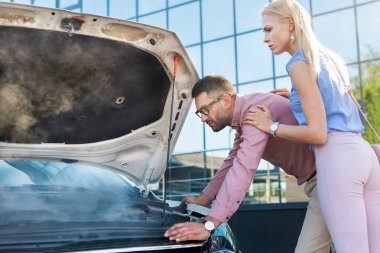 side view of couple looking under car hood of broken car on street clipart