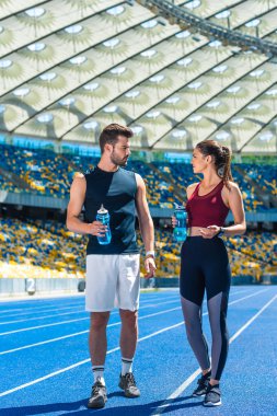 young sportive couple with bottles of water standing on running track at sports stadium and talking clipart