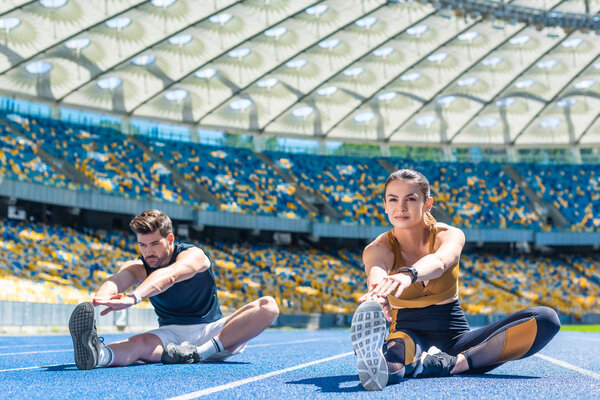 young sportive male and female joggers sitting on running track and stretching at sports stadium