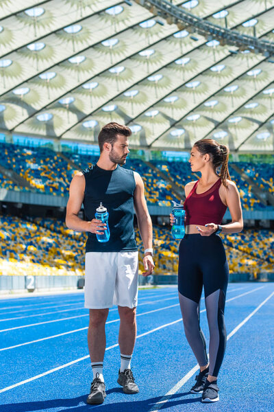young sportive couple with bottles of water standing on running track at sports stadium and talking