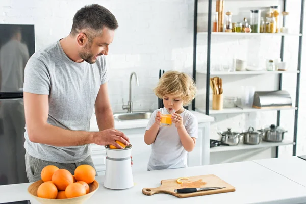 彼の幼い息子がキッチンでジュースを飲みながら絞りでオレンジ ジュースを作って父 — ストック写真
