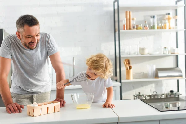 笑顔の父を探してどのようにキッチンで卵黄を泡立て器を舐めている彼の幼い息子  — 無料ストックフォト