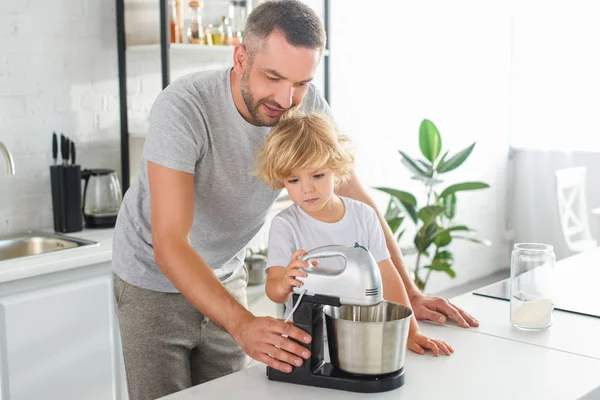Улыбающийся Человек Помогает Сыну Использовать Миксер Приготовления Теста Кухне — стоковое фото