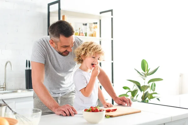 キッチンでイチゴを食べる彼の息子の近くに立って笑顔の父 — ストック写真