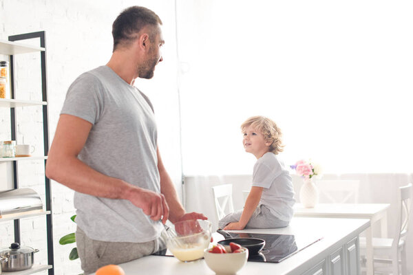 father whisking dough in bowl and talking to son while he sitting on tabletop at kitchen