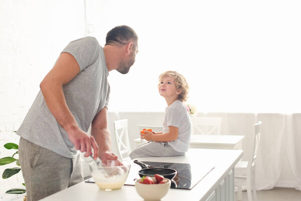 father whisking dough in bowl and talking to smiling son while he sitting on tabletop at kitchen 
