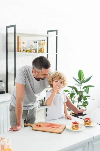 キッチンでの卓上を近く彼の息子の食べるイチゴのパンケーキを見て笑顔の父  — 無料ストックフォト