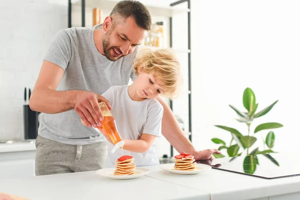 微笑的父亲和儿子浇糖浆在煎饼在厨房 — 图库照片
