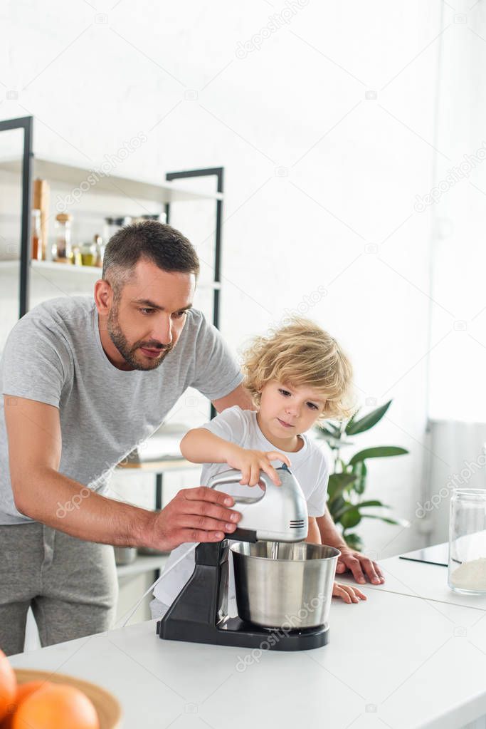 focused man helping his son using mixer for making dough at kitchen 