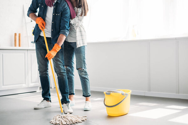 cropped image of boyfriend cleaning floor in kitchen with mop and girlfriend hugging him