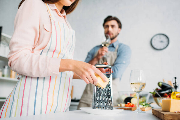 cropped image of girlfriend grating cheese and boyfriend drinking wine in kitchen 