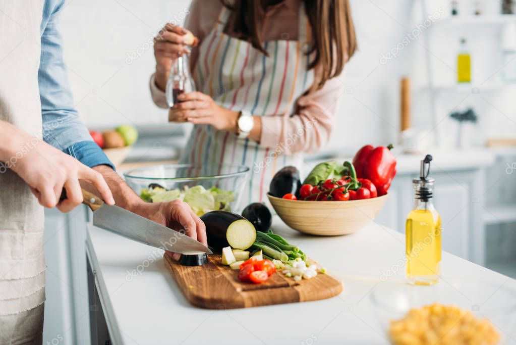 cropped image of boyfriend cutting ingredients and girlfriend adding spices to salad in kitchen