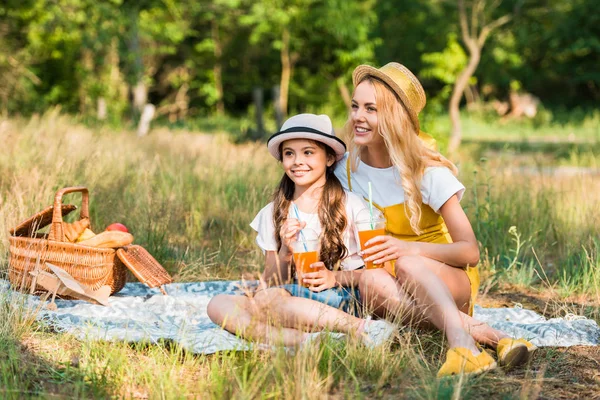 mother and daughter sitting on blanket at picnic and holding glasses of juice