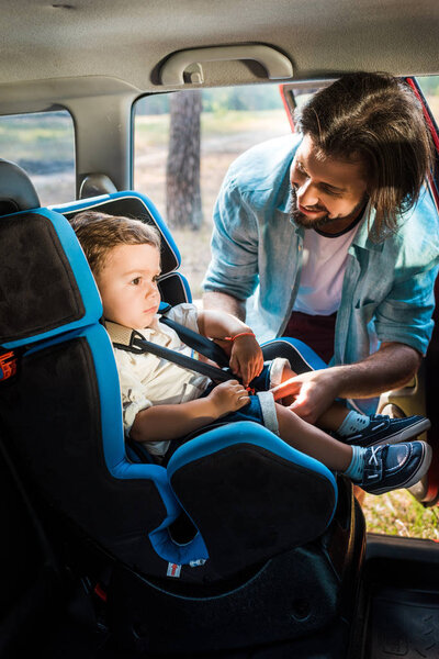 smiling father fastening son in safety seat in car
