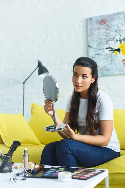 young beautiful woman looking at mirror while sitting on sofa at coffee table with cosmetics at home