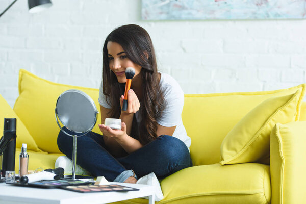 smiling beautiful woman with long hair looking at mirror while applying powder on sofa at home