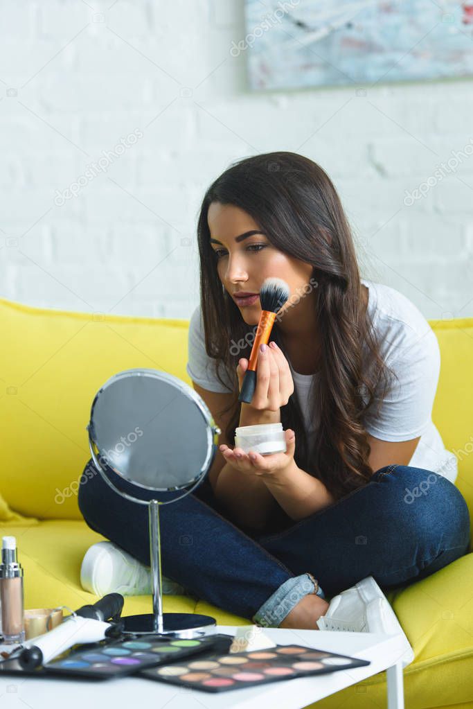 portrait of beautiful woman with long hair looking at mirror while applying powder on sofa at home