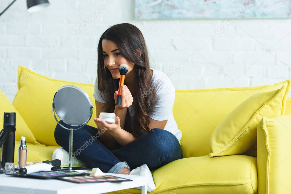 smiling beautiful woman with long hair looking at mirror while applying powder on sofa at home