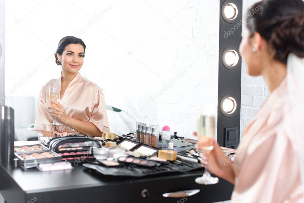 mirror reflection of smiling bride in silk bathrobe and veil with glass of champagne sitting at tabletop with cosmetics