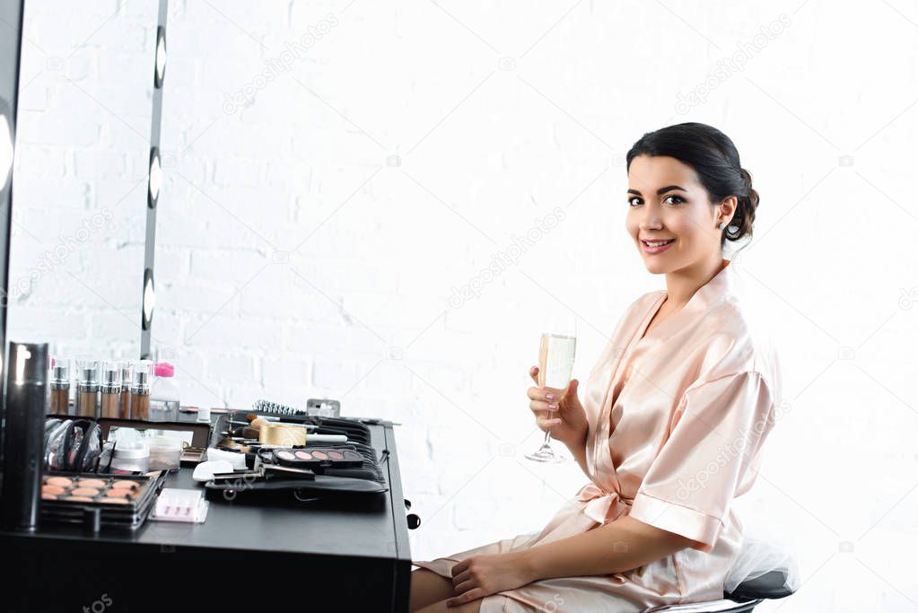 side view of smiling bride in silk bathrobe and veil with glass of champagne sitting at tabletop with cosmetics
