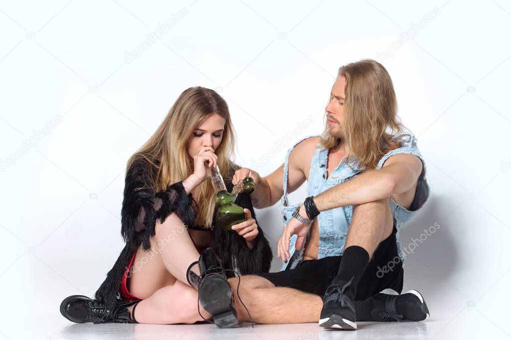 young addicted couple sitting on floor and smoking water pipe of cannabis on white