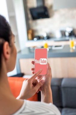 selective focus of woman using smartphone with youtube logo on screen in kitchen clipart