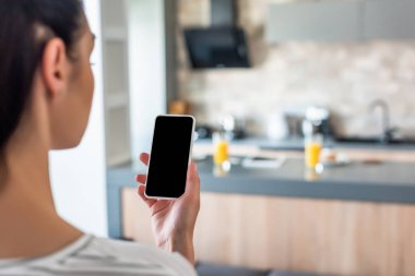 selective focus of woman holding smartphone with blank screen in hand in kitchen clipart