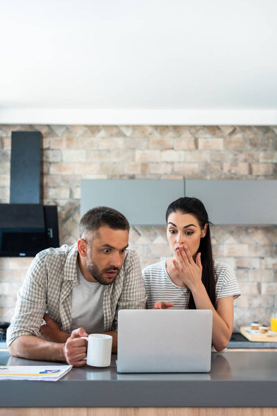 portrait of shocked married couple looking at laptop screen together at counter in kitchen