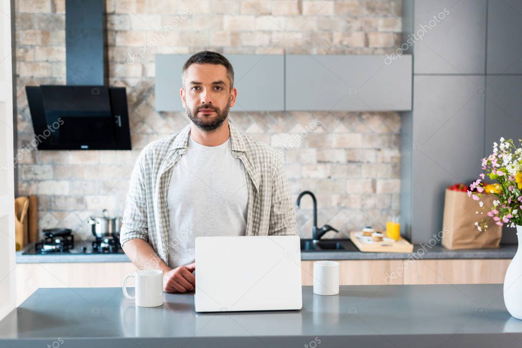 portrait of bearded man standing at counter with laptop and cups of coffee and looking at camera in kitchen