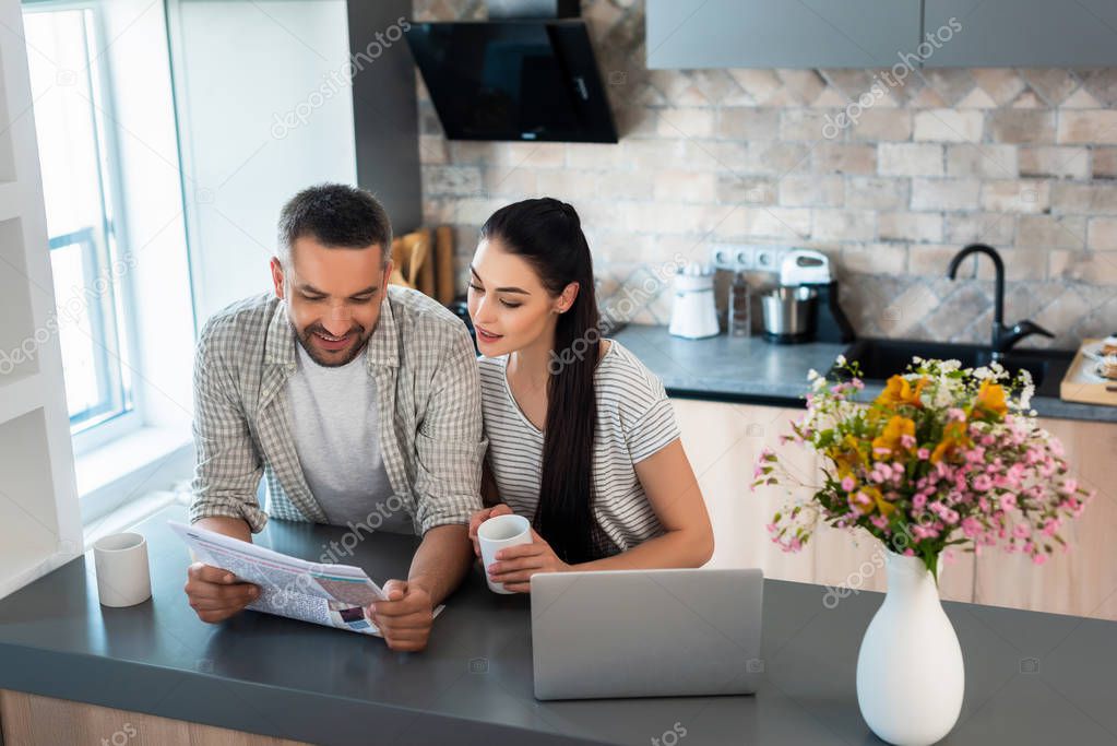Portrait of smiling married couple reading newspaper together at counter with laptop in kitchen
