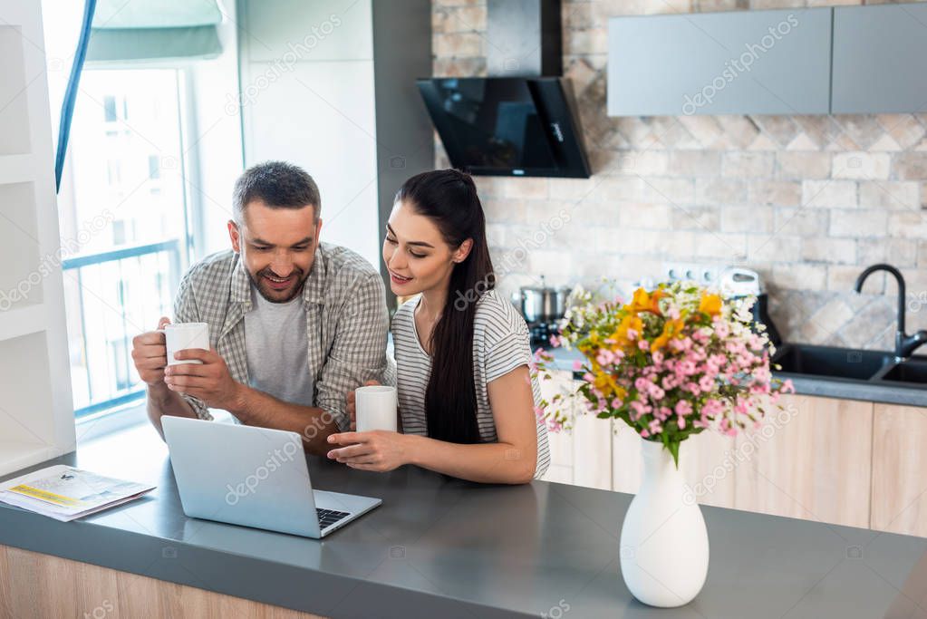 smiling married couple looking at laptop screen together at counter in kitchen