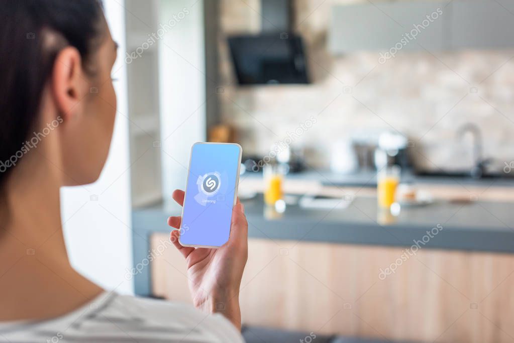 Selective focus of woman holding smartphone with shazam logo on screen in kitchen