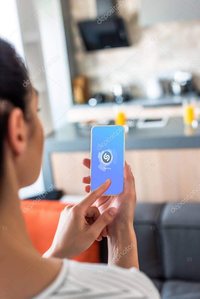 Selective focus of woman using smartphone with shazam logo in kitchen