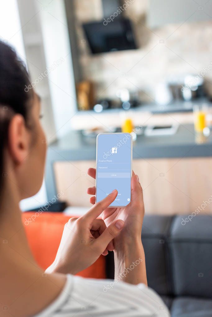 Selective focus of woman using smartphone with facebook logo in kitchen