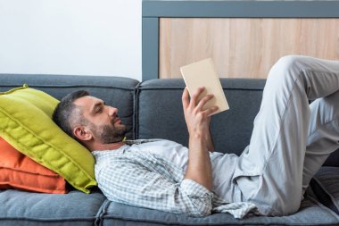 side view of bearded man reading book while lying on couch at home clipart